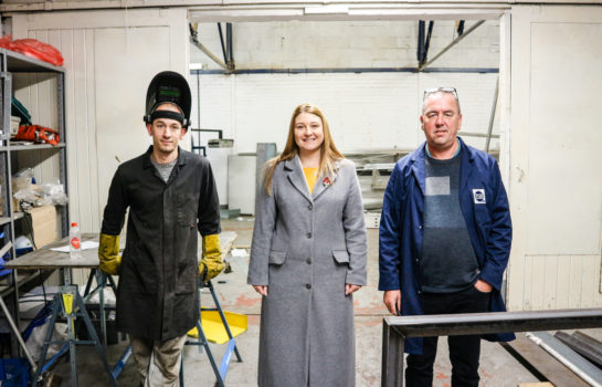 Two men and a woman standing in a workshop, featuring E-Factor Properties.