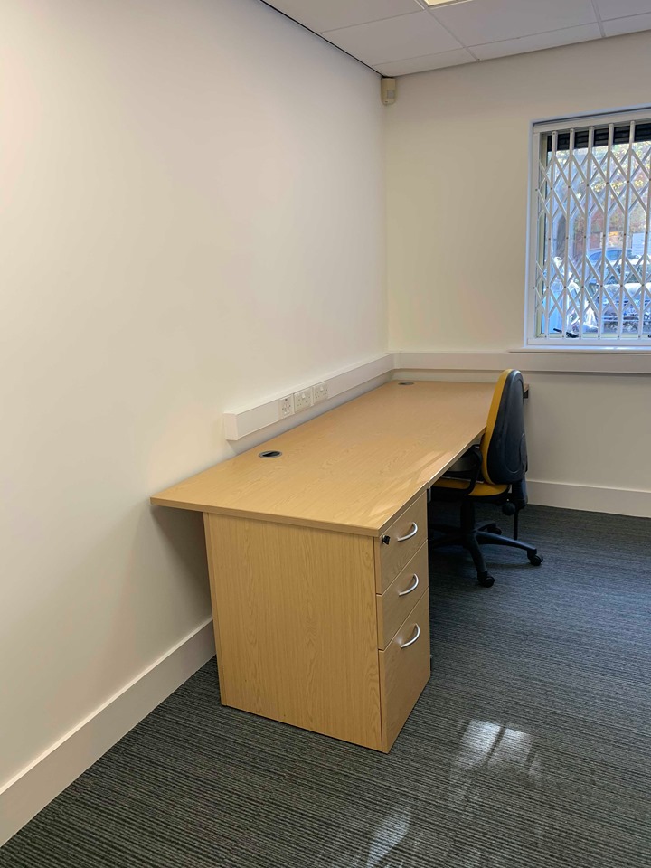 An empty office corner on Dudley Street features a wooden desk with three drawers, an office chair, and a window with a security grate. The white wall contrasts with the gray carpet, creating a professional atmosphere perfect for your next venture at Business Hive in Grimsby. An empty office corner on Dudley Street features a wooden desk with three drawers, an office chair, and a window with a security grate. The white wall contrasts with the gray carpet, creating a professional atmosphere perfect for your next venture at Business Hive in Grimsby.