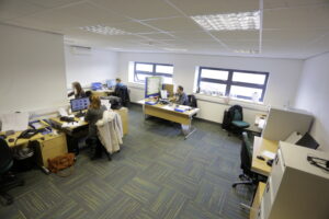 A modern office in F4 Enterprise Village, Grimsby with five people working at desks, each using computers. The room has multiple windows allowing natural light, and there are filing cabinets and office supplies around.