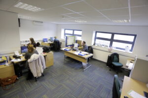 Four people work at desks in an office with large windows overlooking Enterprise Village. Two people are using computers, one is holding papers, and another is writing. The carpeted floor and overhead lighting create a cozy atmosphere in their Grimsby location.