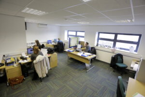 An office space with four people working at desks. The room, located near Prince Albert Gardens, has white walls, multiple windows, and desks equipped with computers and office supplies.