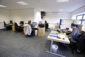 Four people work at desks in a small office space with computers, paperwork, and office supplies. Some desks have personal items like coats and bags. The office, located in F4 Enterprise Village near Grimsby, features a blue carpet and white walls.