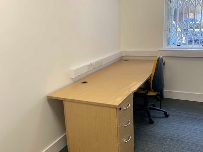 An empty office corner on Dudley Street features a wooden desk with three drawers, an office chair, and a window with a security grate. The white wall contrasts with the gray carpet, creating a professional atmosphere perfect for your next venture at Business Hive in Grimsby.